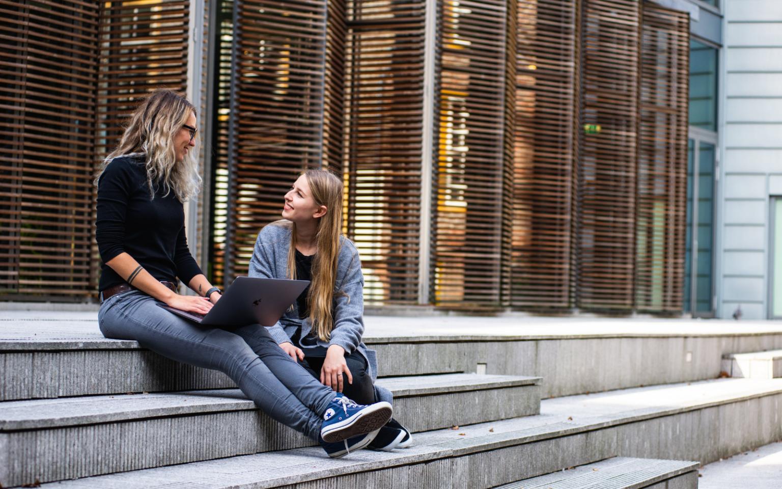 A woman asking a another woman how do I become a recruiter as they sit on the steps outside of a building together